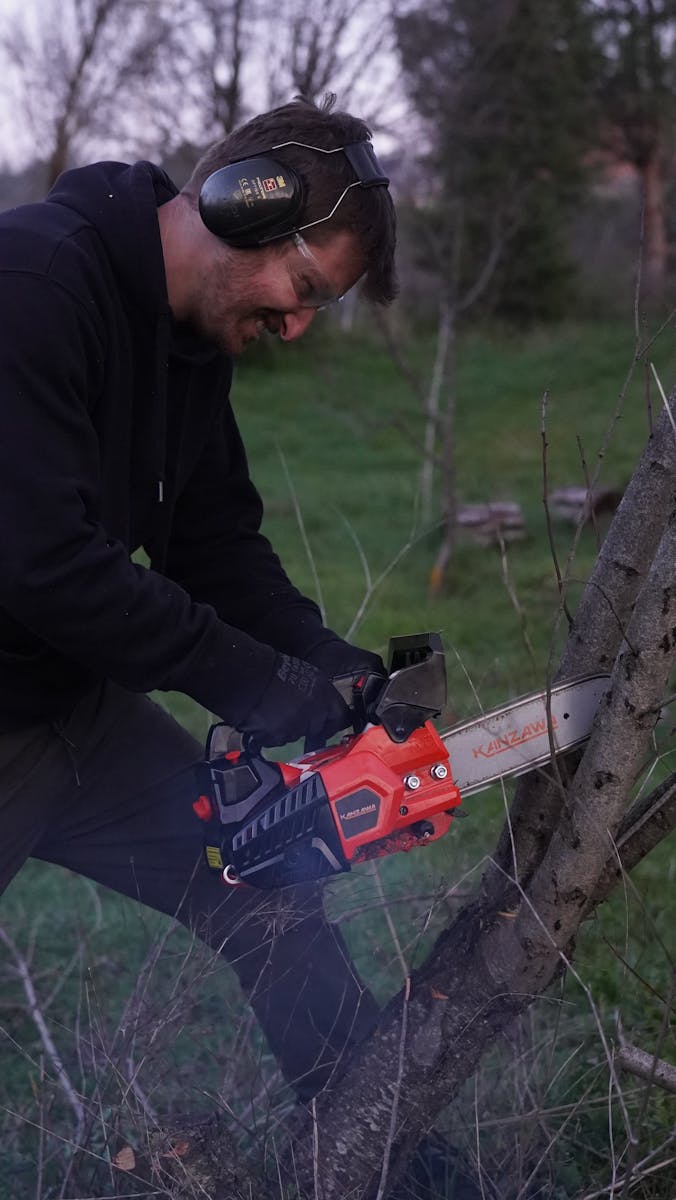 Man in protective gear using a chainsaw to cut a tree outdoors.